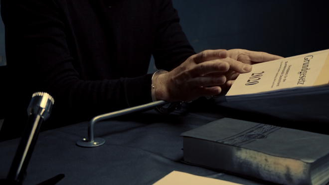 A close up image of a man handcuffed to a table, you cannot see his face. He is holding up a book with the title &ldquo;Grundgesetz&rdquo;. Beside him on the table is what looks like a bible. In the foreground of the shot is a plastic looking microphone. The man&rsquo;s hands are hancuffed to a towel rack which is not screwed into the table.