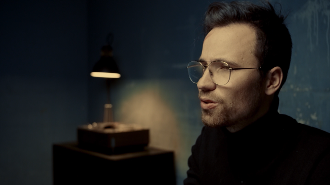 A profile shot of a man sitting in a dimly lit room, evidently talking to someone across from where he is sitting. The walls are a greyish colour. In the corner of the room, a small sits atop a table illuminates some kind of electronic device.