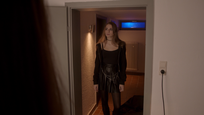 An image of a woman standing in a doorway of a hotel room, facing forward and staring blankly off-camera. She is wearing all black, including a choker, hoody, tank top, pleated wet-look skirt, tights, and boots.