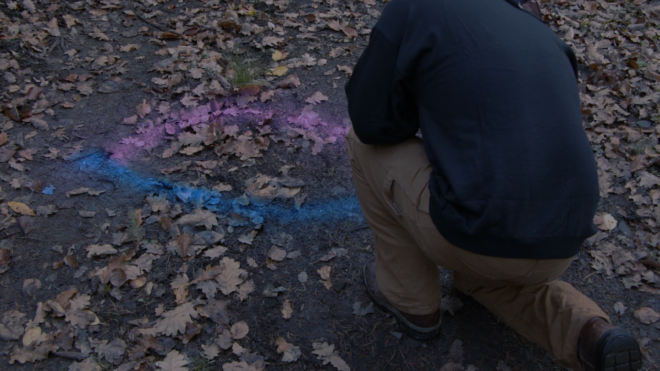 A man crouching on a dirt road in the forest, looking down and away from the camera. On the ground part of a Jesus fish symbol has been spray painted into the ground. The top half is pink while the bottom half is blue.
