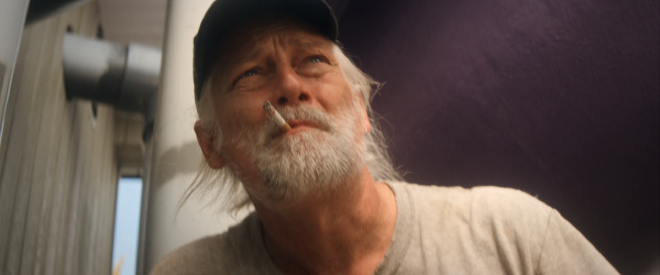 A closeup shot of middle to older age man with medium length grey hair and facial hair. He is wearing a hat and smoking a cigarette while looking a little apprehensively in the distance, off camera.