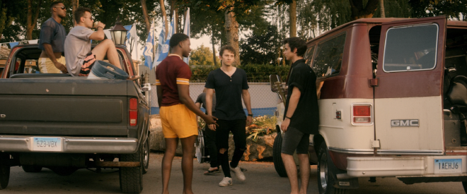 Several male students are standing outside their parked cars and talking. On the right is a couple band members and their van. On the left is a athlete in uniform. To the left of the athlete, several more sit in the back of jacked up truck, drinking beer.
