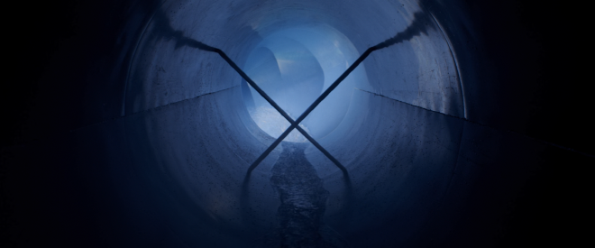 An image taken with the camera pointing directly down the waterslide. It is dark, but there is light at the end of the tunnel. Partway down, large metal razor blades are inserted into the slide, forming an X shape.