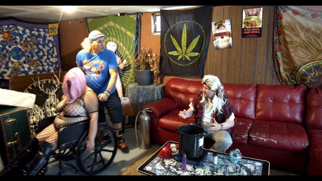 An image taken in a basement that would be stereotypical for a so-called redneck. One man sits on a couch, wearing an awful looking wig. In front of him, on a coffee table, is a pot, portable burner, and various equipment including goggles, a dildo, funnel, and a spice shaker. To the left of him, another man, also wearing a terrible wig, is wheeling in a woman in a wheelchair. She appears to be unconscious, and is wearing a fishnet body suit, knee high boots, and a pink wig.