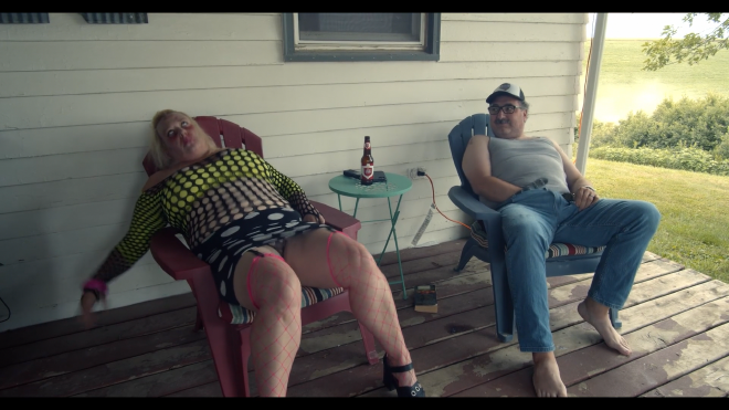 An image of two somewhat overweight people, a man and a woman, sitting on a porch. The man has his hands down his pants and looks excited. Meanwhile, the women looks to convulsing, and is wearing clothing that would typically be associated with a sex worker. She has some blood and nasty looking makeup around and on her face.