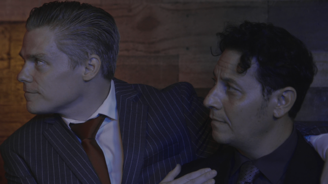 An image of two men standing in front of wood-panelled backdrop. They are both wearing suits, and one man has an around the other. They are both looking off-camera. The backdrop is lit with a neon green LED hue.