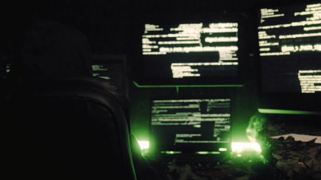A close up image of a hooded figure sitting a computer desk. The scene is dimly lit with flourescent green, and there are mutiple monitors in the background. The computer equipment on the desk is wrapped in camoflouge webbing.