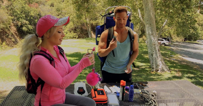 An image of the main character, Barbi, and her boyfriend, Jake, outdoors. Barbi is wearing all pink, and sitting on a picnic table while looking intently at the cell phone in her hands. Meanwhile, Jake is carrying a large hiking backpack and standing at the side of the table. On the table he has arranged an array of different camping/hiking supplies and is currently examining one of them.