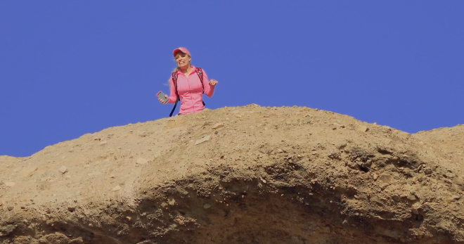 An image taken from below, with Barbi standing at the edge of cliff and looking down over the edge. She looks distressed, phone in hand, like she just did something really embarrassing.