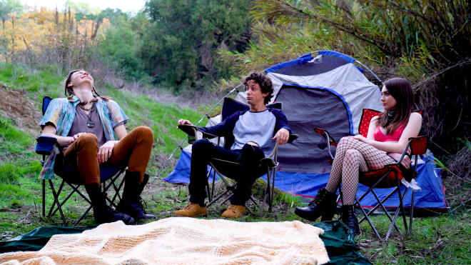 A group of 3 people sit in camping chairs in a forest. The two male characters are mostly dressed appropriately for the wilderness, while the female character is not, and is wearing heeled boots. Their chairs are spread in front of a blanket on the ground, and in the background is a pitched tent.