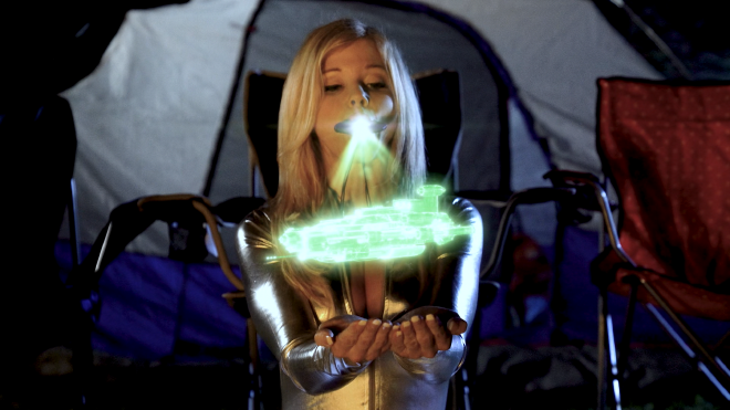 The character Sheera with her large quartz crystal in her mouth, sitting in front of a tent and some camping chairs in the dark. From the quartz, a green holographic depiction of an alien spaceship is displayed.