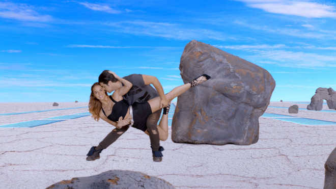 A woman and man holding a dance pose. They are in front of computer generated landscape compose of several large rocky structures on an otherwise clear flat ground. There are blue rectangles on the ground in the distance, but it&rsquo;s not clear what they are.