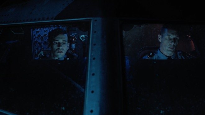 An image taken from outside a commercial airliner, looking straight through the front windows of the cockpit. It&rsquo;s dark and rainy outside. Inside the cockpit are two men, strapped in and wearing pilot&rsquo;s uniforms.