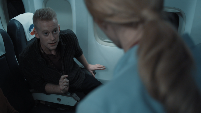 An agitated looking man is seated and buckled up on a plane. He is leaning out of his chair, looking intently at a woman in the foreground of the shot, with her back turned to the camera. She is wearing a steward uniform.
