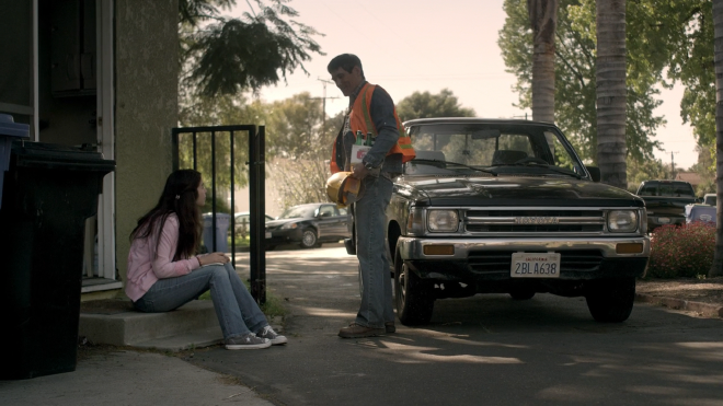 The character Kayla sitting outside the entryway to their green house, homework in hand. Her father, Ken, is standing in front of his truck, wearing a orange reflective vest and holding a 6-pack of beer and a construction helmet.