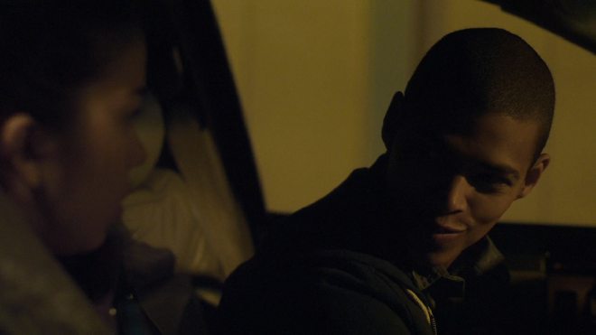 A close up shot through the side window of a car. Kayla is leaning back in her seat and looking at Treyshawn, while he is leaning forward and talking to her. It is very dark outside.