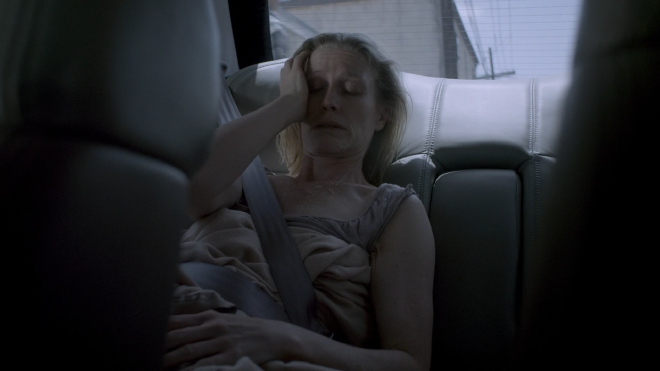An image of the mother Angie, sitting in the back seat of car. She looks very unhealthy, strung out, and has vomit down her front.