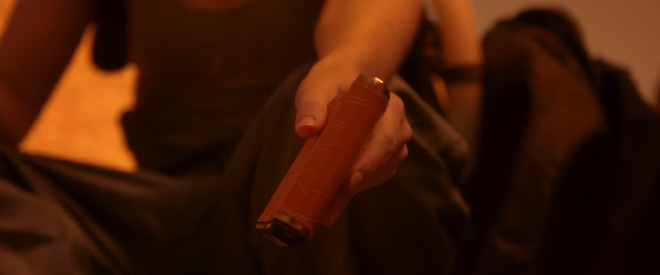 A close-up shot of a character holding a flask out. They are sitting on the ground in the desert, with a backpack beside them. The image is tinted red, as they are on Dune.