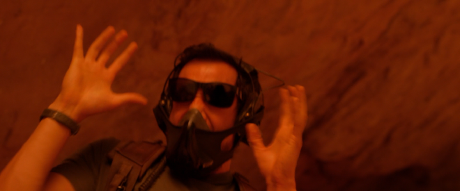 A man wearing sunglasses and breathing mask like at a construction site, along with some military gear. He has raised his hands in panic and looks scared. The entire shot is very red, and set against a red rock backdrop.