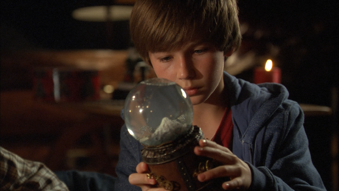 A young boy is sitting on the floor. In his hands is an intricate snow globe, which he is staring intently at. The globe has a large, heavy looking base with clockwork-style gears around it.