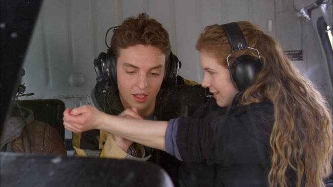 A young woman sits in a seat of helicopter, wearing a headset. Behind her is a young man, who is leaning forward and signing her arm with a marker. She looks delighted.