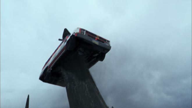 An image, taken from below, of a car impaled with a huge, rock-like spike. The car is lifted way off the ground.