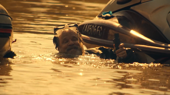 An image of a middle-aged man in scuba gear, mostly submerged in water. He is holding a cable in one hand, which is stick out of the water, and holding onto the side of a small boat.