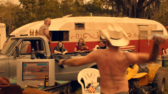 An image of several people leaning against or standing against an old pickup truck. The bed of the truck is lined, and a duck is sitting in it. Strewn about the scene are bottles of beer and various trash. In the background is a beat-up looking trailer, and in the foreground a shirtless man in a cowboy hat is in the process of throwing a bottle of beer at someone.