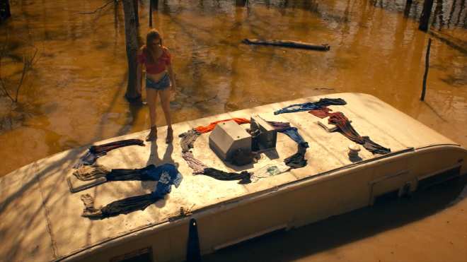 An image of a women standing on the roof of a mostly submerged trailer in muddy looking water. She has arranged various pieces of wet clothing to spell out SOT.