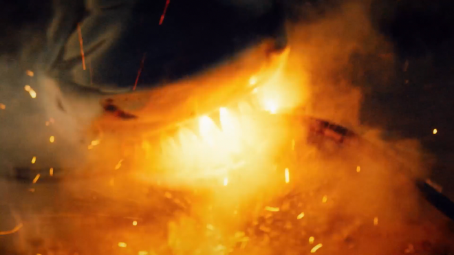 An image of shark, partially submerged in water, biting down on an electrified cable. It looks like the shark is receiving a strong electric shock and perhaps catching on fire. There is a very bright orange glow coming from it’s mouth, with smoke billowing out.
