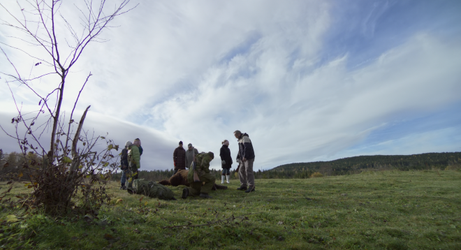 An image of a group of people, several hunters, gathered around a dead bear out in a field. The green, grassy field takes up the lower third of the shot, while blue cloudy sky takes up the top third. Mild, rolling hills are seen in the distance.
