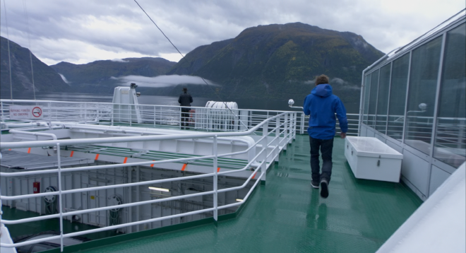 An image taken on a ferry, which has a green deck and white railing. Hans is standing on the far side of the ferry, looking into the water. One of the students walks toward him, back to the camera. In the background you can see mountains, water, sky and clouds.