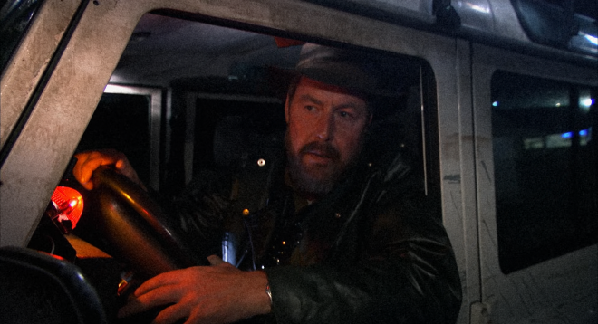 A man sits in the driver&rsquo;s seat of a reinforced jeep at night, one arm outside the open window, one hand on the wheel. He is wearing a hat and black jacket. The jeep is silvery-white in colour, heavily scratched up and adorned with various tactical looking attachments.
