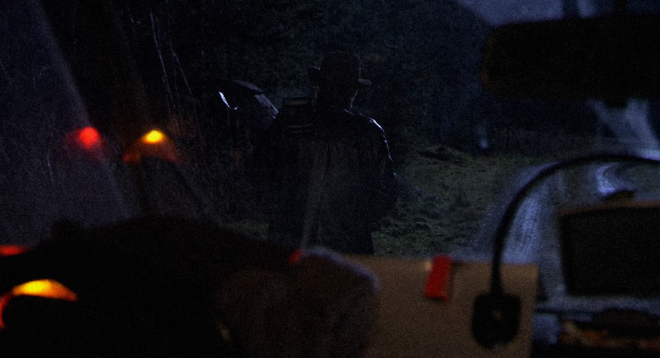An image taken from inside a vehicle at night, looking through the windshield. It is very dark and grainy. Hans has his back to the camera, and is holding a spotlight attached to a stick while he peers into the forest.